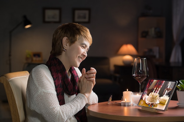 Donna di mezza età asiatica che celebra il compleanno virtuale tramite videochiamata a casa durante l'allontanamento sociale