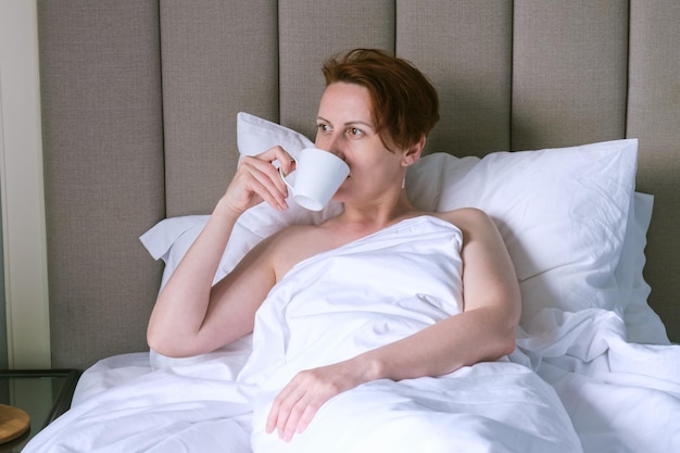 Donna di mezza età beve caffè a letto La donna riposa in un hotel e si rilassa