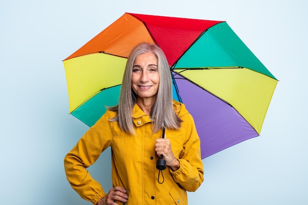 Donna di mezza età capelli grigi sorridente felicemente con una mano sull'anca e sicura di sé. concetto di ombrello e pioggia