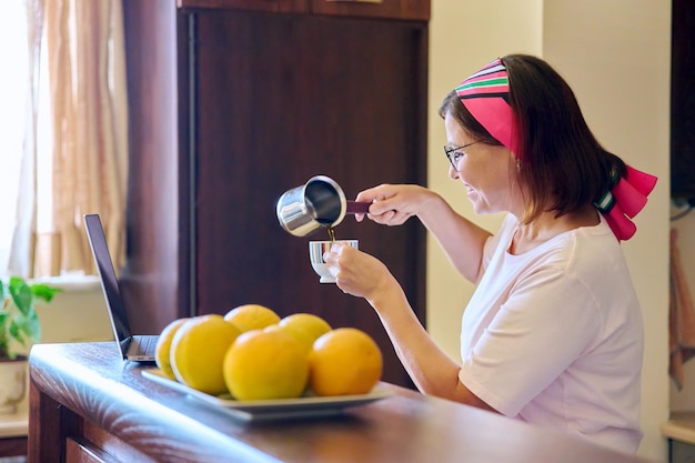 Donna di mezza età che beve caffè parlando in videochiamata utilizzando la cucina del computer portatile a casa