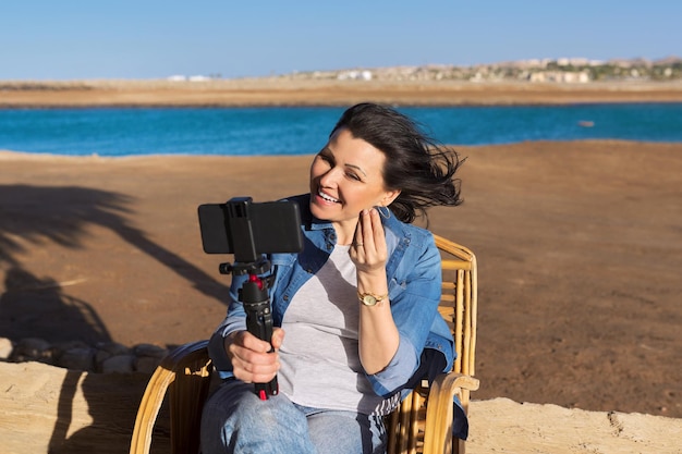 Donna di mezza età che guarda la webcam dello smartphone che parla di registrazione di video sulla spiaggia sabbiosa