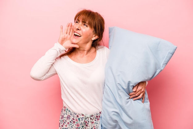 Donna di mezza età che indossa un pigiama isolato su sfondo rosa che grida e tiene il palmo vicino alla bocca aperta