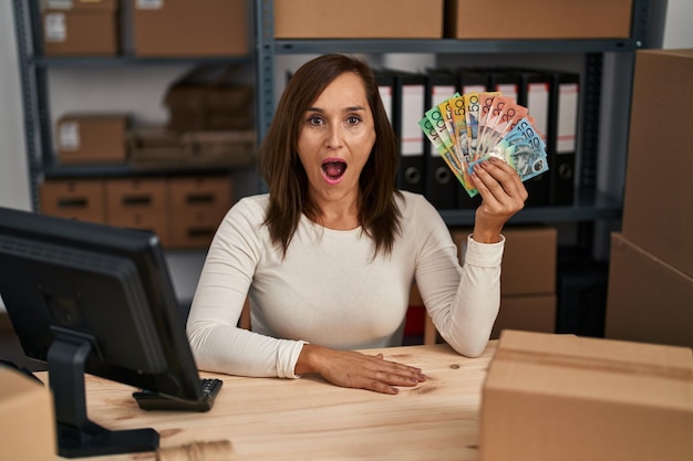 Donna di mezza età che lavora presso un e-commerce di piccole imprese con dollari australiani spaventati e stupiti con la bocca aperta per il viso incredulo a sorpresa