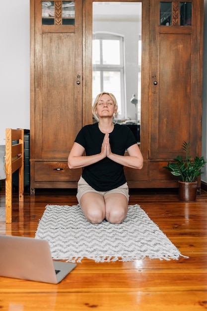 Donna di mezza età che media e fa yoga a casa con la lezione online del laptop