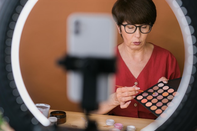 Donna di mezza età che parla di cosmetici con ombretti per il trucco e palette di fard e pennelli mentre registra il suo video Donna matura che fa video per il suo blog sui cosmetici