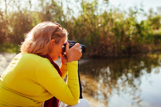 Donna di mezza età che prende le immagini sulla macchina fotografica che si siede dalla sponda del fiume di autunno.