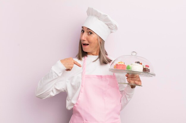 Donna di mezza età che sembra scioccata e sorpresa con la bocca spalancata che punta al concetto di panettiere e torte