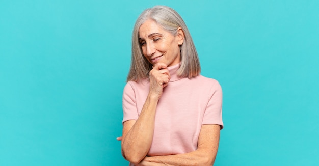 Donna di mezza età che sorride con un'espressione felice e sicura con la mano sul mento, chiedendosi e guardando di lato