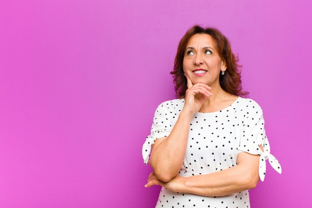 Donna di mezza età che sorride felicemente e sognare ad occhi aperti o dubitare, guardando al lato contro il muro viola