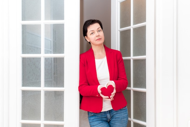 Donna di mezza età che tiene nelle sue mani un grande cuore rosso di San Valentino vicino alla porta bianca.