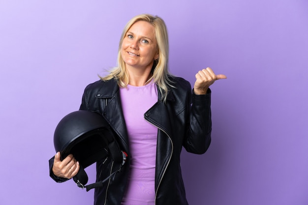 Donna di mezza età che tiene un casco da motociclista isolato sulla parete viola che punta di lato per presentare un prodotto