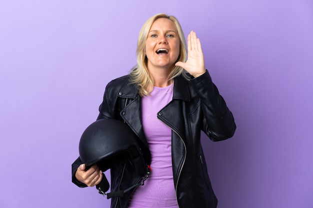 Donna di mezza età che tiene un casco del motociclo isolato sul muro viola che grida con la bocca spalancata