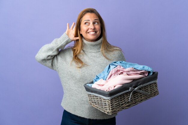 Donna di mezza età che tiene un cesto di vestiti isolato sulla parete viola ascoltando qualcosa mettendo la mano sull'orecchio