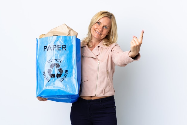 Donna di mezza età che tiene un sacchetto di riciclaggio pieno di carta da riciclare isolato sul muro bianco facendo gesto in arrivo