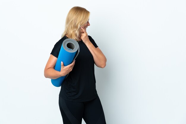 Donna di mezza età che va alle lezioni di yoga mentre si tiene un tappetino isolato sul muro bianco pensando un'idea e guardando il lato