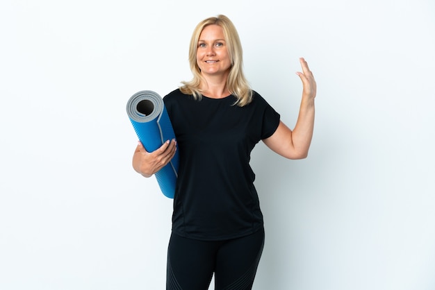 Donna di mezza età che va alle lezioni di yoga mentre si tiene una stuoia isolata sul muro bianco che saluta con la mano con l'espressione felice