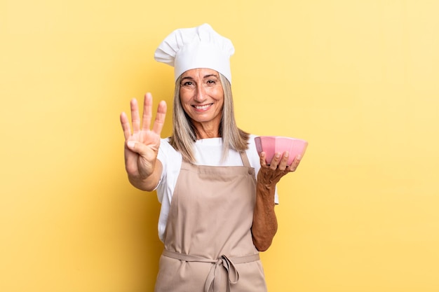 Donna di mezza età chef sorridente e amichevole che mostra il numero quattro o quarto con la mano il conto alla rovescia e tenendo una pentola vuota