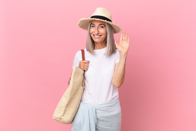 Donna di mezza età con capelli bianchi che sorride felicemente, agitando la mano, dandoti il benvenuto e salutandoti. concetto di estate