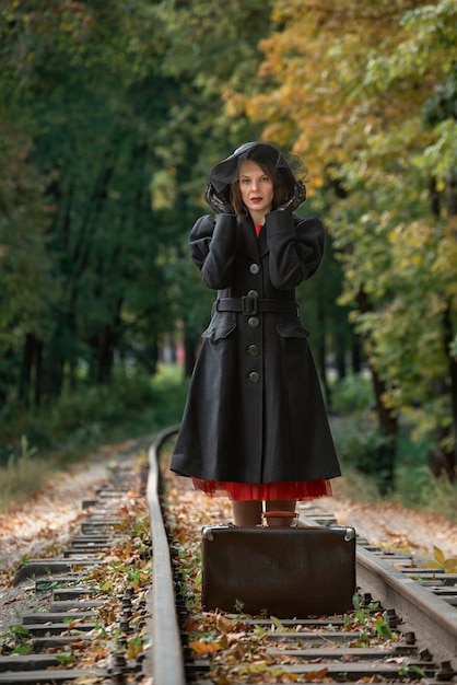 Donna di mezza età con cappello a tesa larga con cappotto nero retrò si trova vicino a una valigia vintage sui binari della ferrovia Cornice verticale