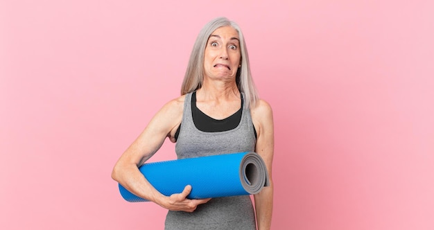 Donna di mezza età con i capelli bianchi che sembra perplessa e confusa e tiene in mano un tappetino da yoga. concetto di fitness