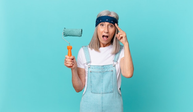 Donna di mezza età con i capelli bianchi che sembra sorpresa, realizzando un nuovo pensiero, idea o concetto con un rullo che dipinge un muro