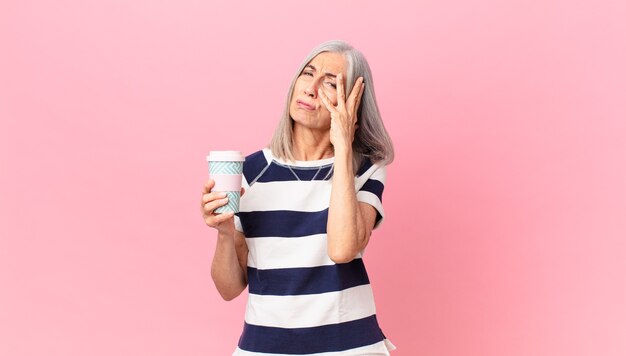 Donna di mezza età con i capelli bianchi che si sente annoiata, frustrata e assonnata dopo una noiosa e con in mano un contenitore di caffè da asporto