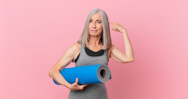 Donna di mezza età con i capelli bianchi che si sente confusa e perplessa, mostra che sei pazzo e tiene in mano un tappetino da yoga. concetto di fitness