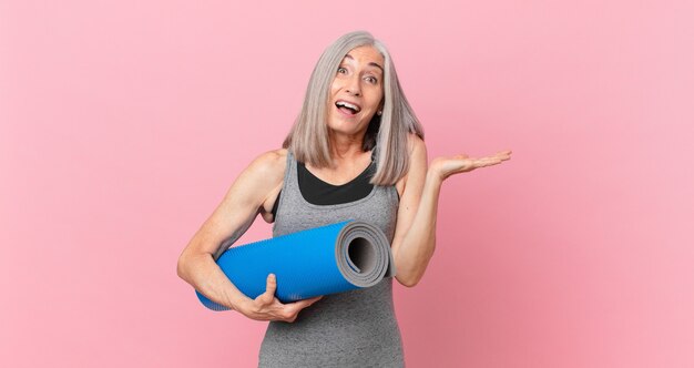 Donna di mezza età con i capelli bianchi che si sente felice e stupita per qualcosa di incredibile e tiene in mano un tappetino da yoga. concetto di fitness