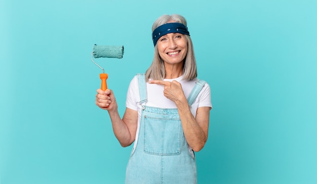 Donna di mezza età con i capelli bianchi che sorride allegramente, si sente felice e indica il lato con un rullo che dipinge un muro