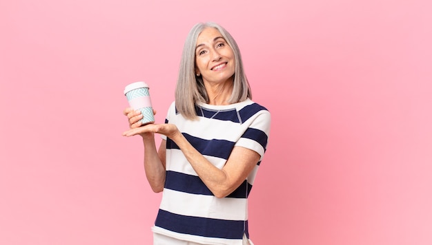 Donna di mezza età con i capelli bianchi che sorride allegramente, si sente felice e mostra un concetto e tiene in mano un contenitore di caffè da asporto