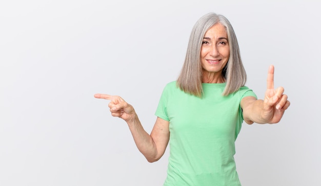 Donna di mezza età con i capelli bianchi che sorride con orgoglio e sicurezza facendo il numero uno e indicando il lato