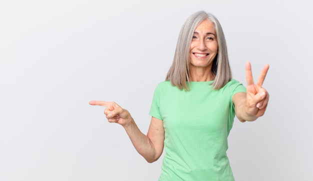 Donna di mezza età con i capelli bianchi che sorride e sembra amichevole, mostra il numero due e indica il lato