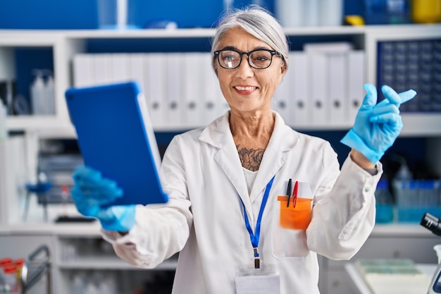 Donna di mezza età con i capelli grigi che lavora al laboratorio dello scienziato facendo una videochiamata sorridendo felice indicando con la mano e il dito di lato