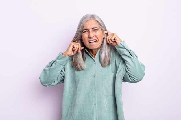 donna di mezza età con i capelli grigi che sembra arrabbiata, stressata e infastidita, coprendo entrambe le orecchie con un rumore assordante, un suono o una musica ad alto volume