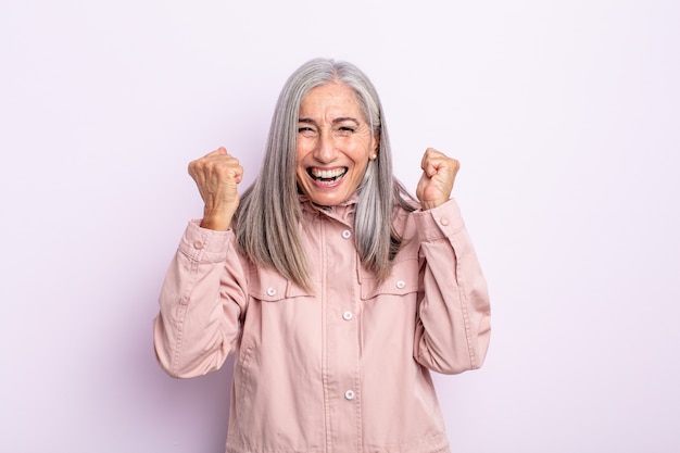 Donna di mezza età con i capelli grigi che si sente felice, sorpresa e orgogliosa, grida e celebra il successo con un grande sorriso