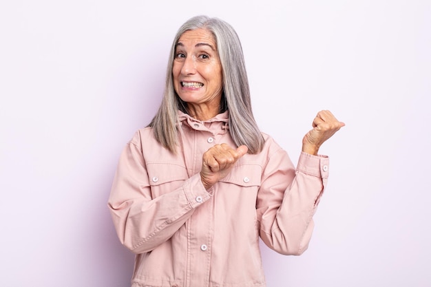 Donna di mezza età con i capelli grigi che sorride allegramente e casualmente indicando lo spazio sul lato, sentendosi felice e soddisfatta