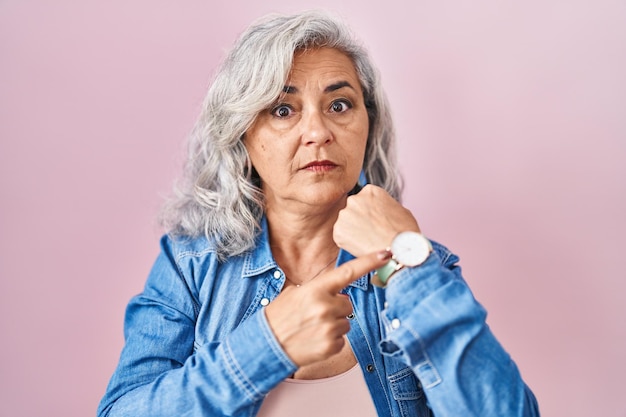 Donna di mezza età con i capelli grigi in piedi su sfondo rosa in fretta che punta a guardare il tempo, impazienza, guardando la telecamera con espressione rilassata