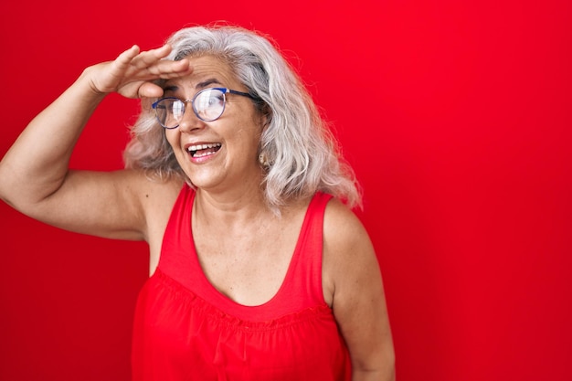 Donna di mezza età con i capelli grigi in piedi su uno sfondo rosso molto felice e sorridente guardando lontano con la mano sopra la testa cercando il concetto