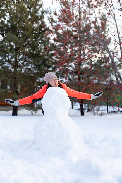 Donna di mezza età con le mani tese e la carota in bocca in piedi dietro il pupazzo di neve incompiuto che distoglie lo sguardo la sera con sorbo e abeti sullo sfondo Tempo invernale in famiglia fuori