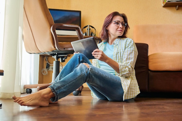 Donna di mezza età con tablet digitale sul posto di lavoro domestico con sfondo del computer