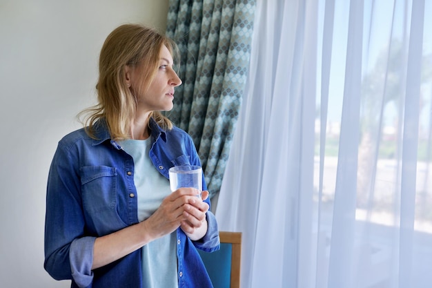 Donna di mezza età con un bicchiere d'acqua in piedi a casa vicino allo spazio della copia della finestra