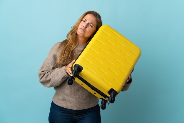 Donna di mezza età del viaggiatore che tiene una valigia isolata sull'azzurro in vacanza con la valigia di viaggio e infelice