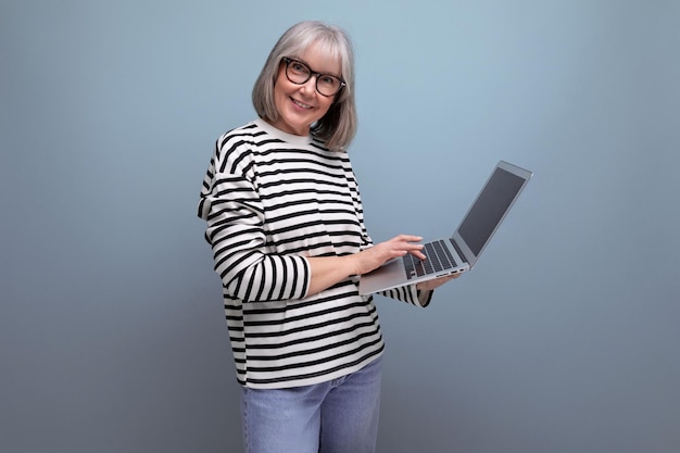 Donna di mezza età di successo con capelli grigi con un computer portatile su uno sfondo luminoso studio con copia