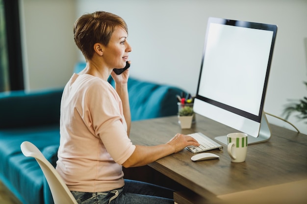 Donna di mezza età freelance che parla su un telefono cellulare mentre lavora al computer in ufficio a casa.