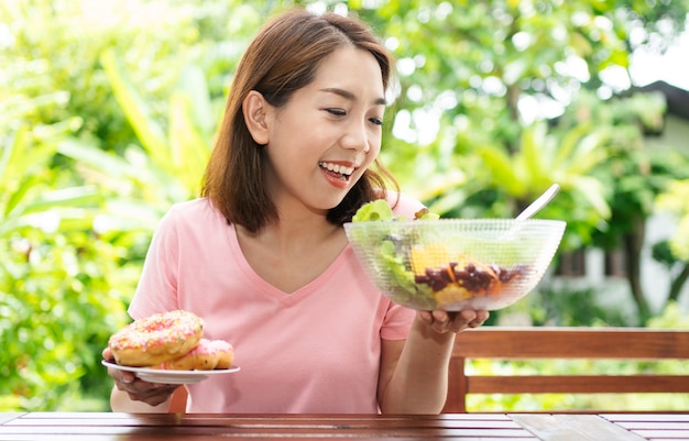 Donna di mezza età in buona salute asiatica che si siede e sceglie tra una ciambella e un'insalata di verdure