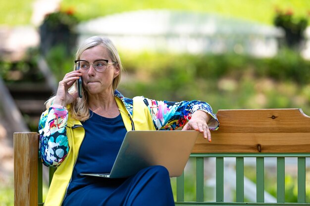 Donna di mezza età in giardino che lavora da casa usando il laptop e parlando al telefono