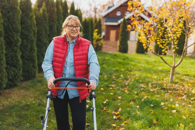 Donna di mezza età in giubbotto rosso con tosaerba sul cortile guardando la fotocamera Giardiniere femminile che lavora in estate o in autunno che taglia l'erba nel cortile Concetto di lavoro di giardinaggio natura