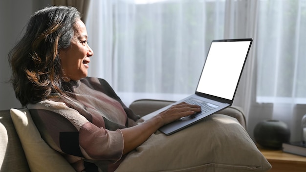 Donna di mezza età sorridente che si rilassa sul divano e utilizza il computer portatile in un luminoso soggiorno moderno