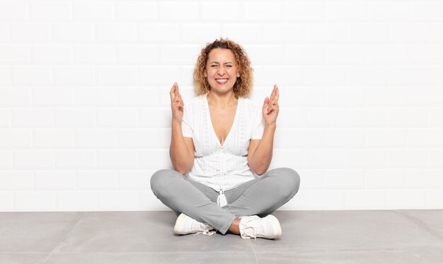 Donna di mezza età sorridente e incrociando ansiosamente entrambe le dita, preoccupata e desiderando o sperando buona fortuna