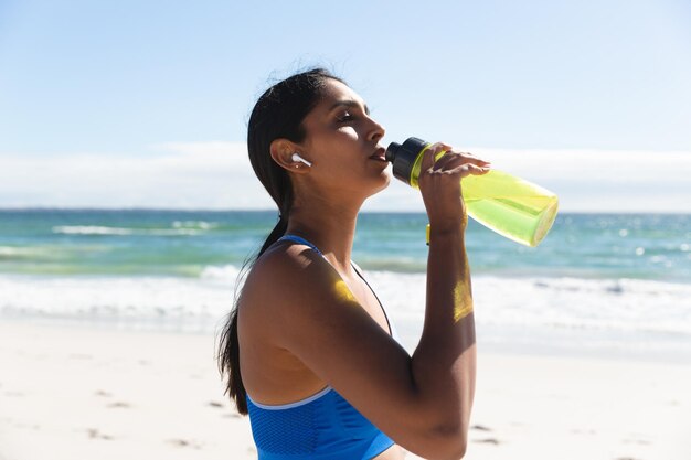 Donna di razza mista che si esercita sulla spiaggia indossando auricolari wireless acqua potabile. sano tempo libero all'aria aperta in riva al mare.